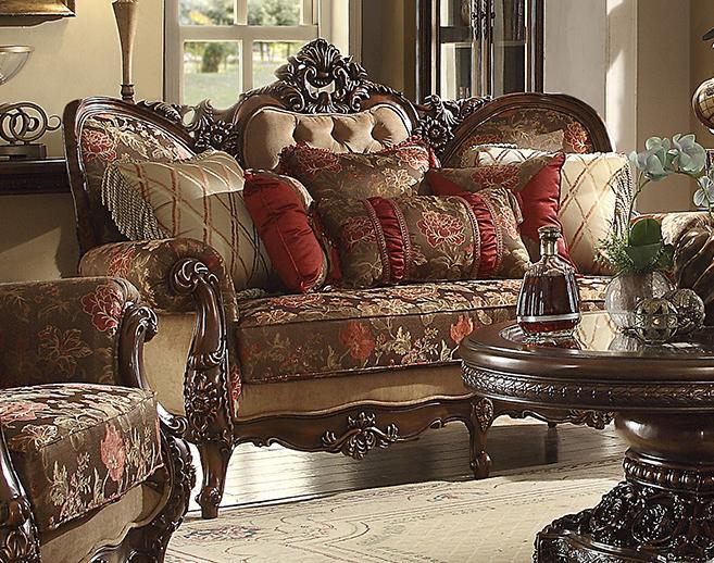 An ornate traditional loveseat sofa with carved dark wood arms and back, upholstered in floral damask fabric with red and beige accents, styled in a classic living room setting with matching pillows and a matching ottoman.